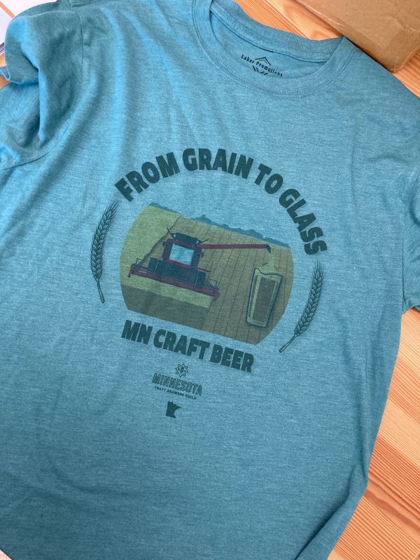 Green shirt with rounded text saying "From Grain to Glass" above an image of a combine harvesting a field; the grain from the unloader is coming out as beer and pouring directly into a pint glass. Below the design are the words "MN Craft Beer" and the MNCBG logo and state outline.